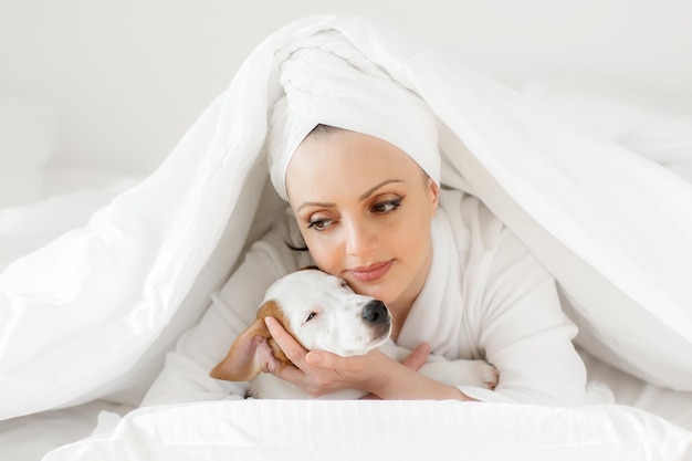 A girl in a white bath towel on her head and a bathrobe in the morning in bed with a dog opens a bir...