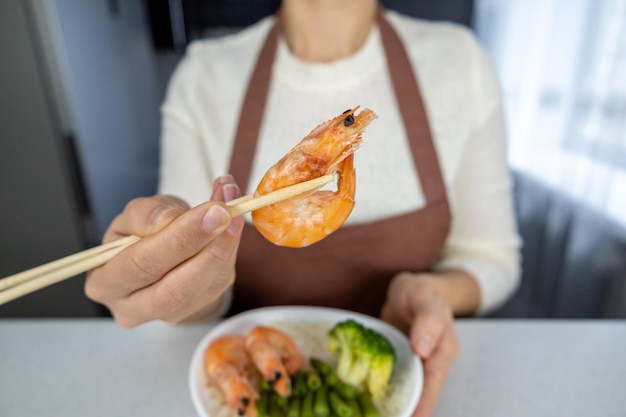 A girl in a white blouse and brown apron holds a large red king prawn with chopsticks On the table is a plate of green beans and broccoli Asian food concept