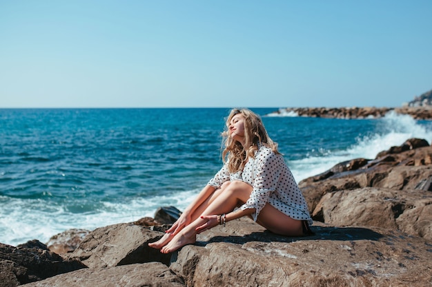 A girl in a white blouse sits on the stones near the sea