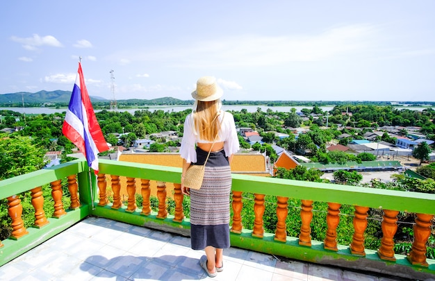 A girl in a white blouse, skirt and hat stands near the Thai flag on the background of the view from the Tiger Cave Temple