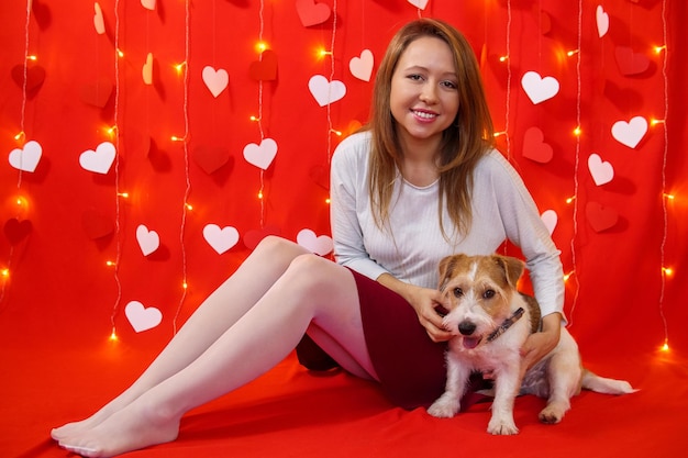 A girl in a white blouse and skirt sits with a dog on a red background with hearts. Valentine's day concept.