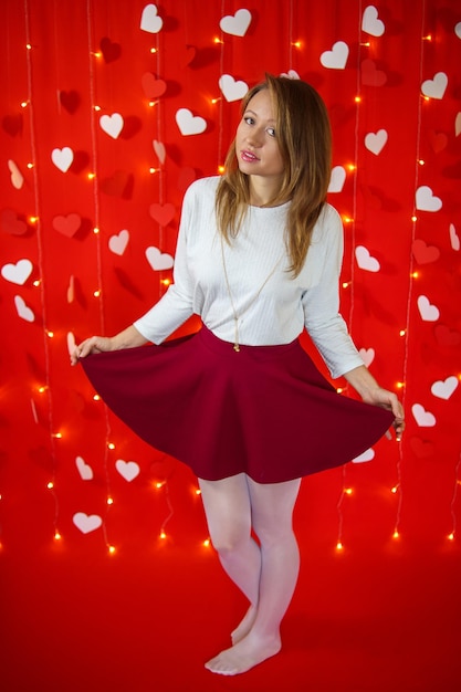 A girl in a white blouse and skirt stands on a red background with hearts. Valentine's day concept.