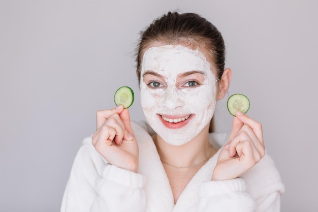 Ragazza in maschera cosmetica bianca sul viso con fetta di cetriolo in mano