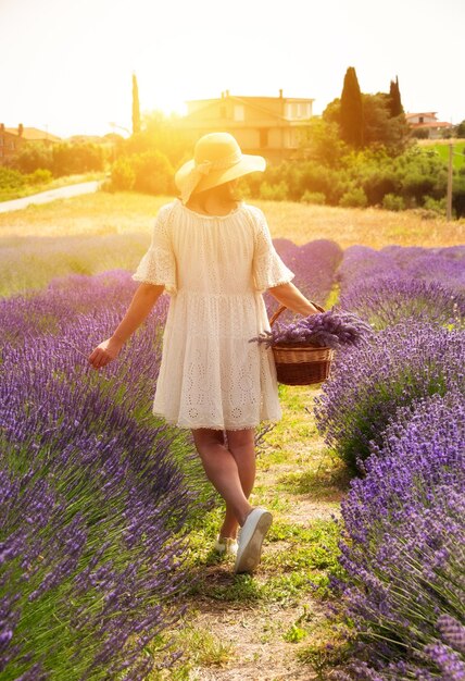 Foto ragazza in abito bianco e cappello con cesto che cammina nel campo di lavanda