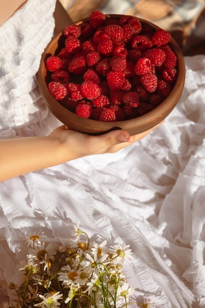 A girl in a white dress holds a clay plate with red ripe raspberries next to a bouquet of daisies