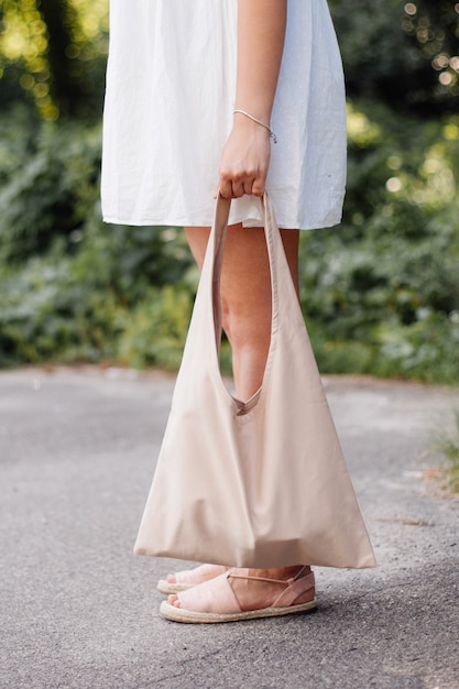 Girl in a white dress holds in her hand a fabric beige bag\
against a surface of greenery