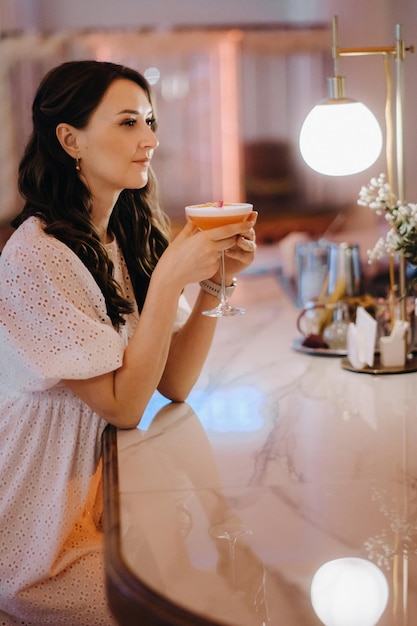 A girl in a white dress is sitting at the bar in a cafe and drinking a cocktail