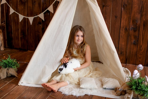 a girl in a white dress is sitting in a tent wigwam with a rabbit, Easter theme