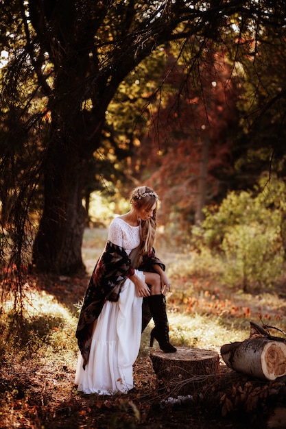 Foto ragazza in abito bianco e poncho in stivali estivi al tramonto nel bosco