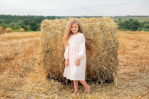 A girl in a white dress stands in front of a stack of hay.