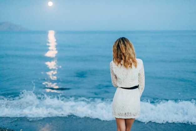Девушка в белом платье стоит спиной на пляже у моря