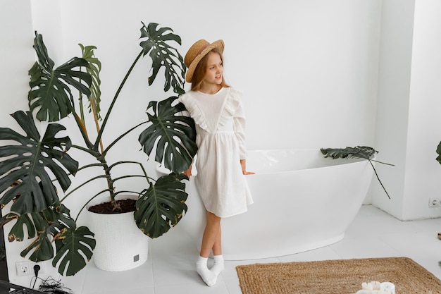 a girl in a white dress and a straw hat in a room with plants and a bathroom