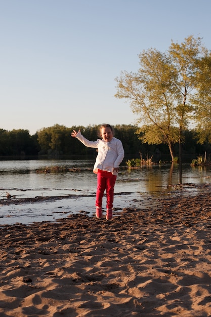 A girl in a white jacket and pink pants jumps on the sand on the beach on the river Bank during sunset.