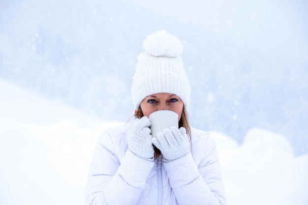 A girl in a white jacket and a white hat is holding a cup of tea in her hands during a snowfall against the background of beautiful snowdrifts