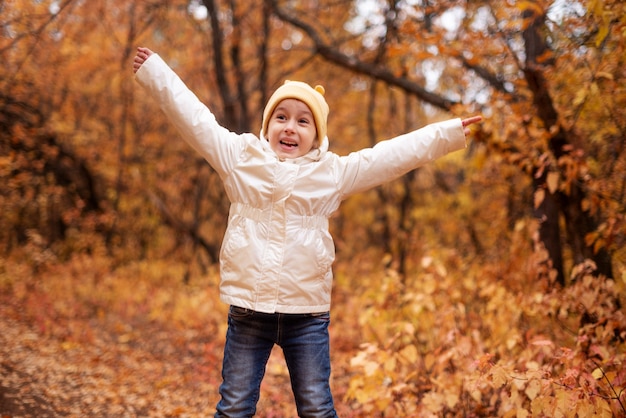 Una ragazza con una giacca bianca e un cappello giallo, un bambino in età prescolare sparge felicemente foglie gialle nella foresta colorata autunnale. il concetto di riposo nella foresta in autunno.