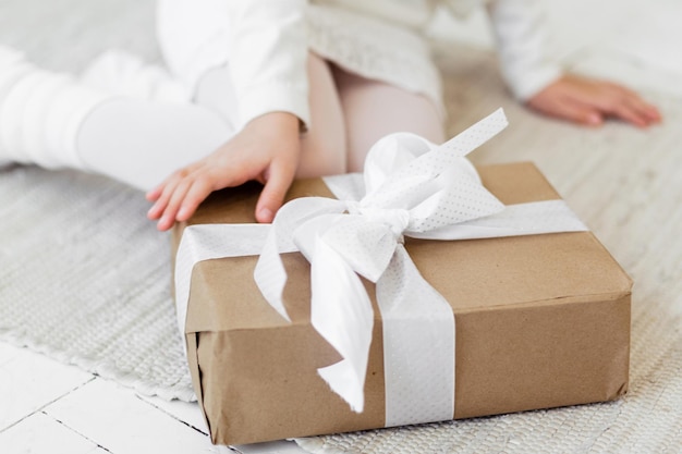 A girl in a white knitted dress sits on the floor and is about to open a gift in craft packaging with a white ribbon