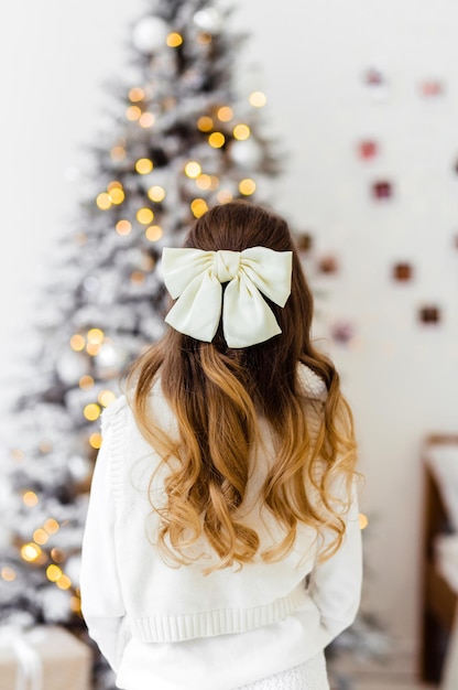 A girl in a white knitted dress with hairpins in the form of a\
bow stands with her back to the camera and looks at the christmas\
tree