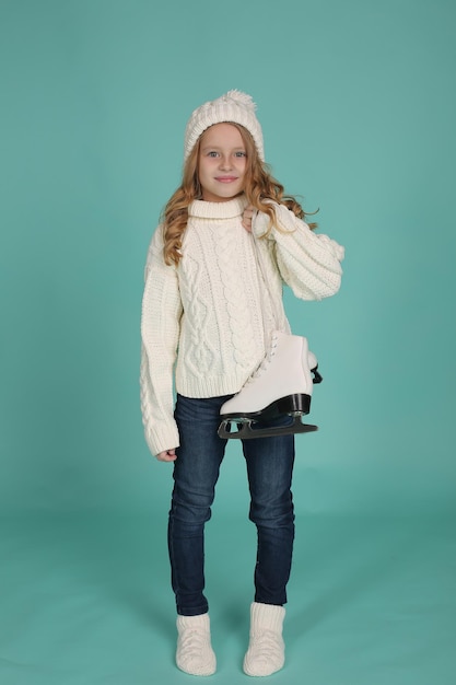 a girl in a white knitted sweater and a white hat holds white figure skating skates in her hands