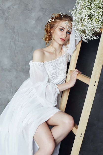 Girl white light dress and curly hair, portrait of woman with flowers at home near the window, purity and innocence. Curly blonde romantic look, beautiful eyes. White wildflowers in hands