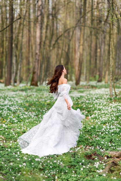 Ragazza in un abito lungo bianco sta girando in una radura con fiori una giovane donna nella foresta di primavera