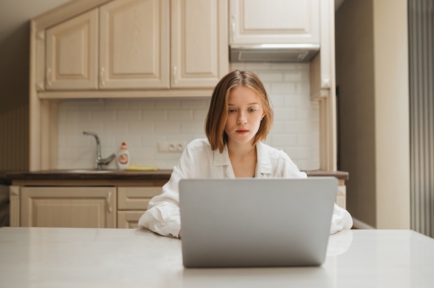 Girl in a white shirt uses a laptop at home in the kitchen