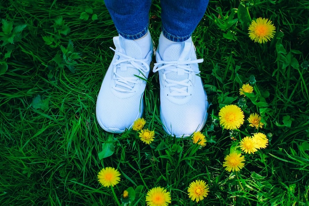 Girl in white sneakers stand near the Yellow dandelions in the green spring grass Sunny summer day Relax and travel concept