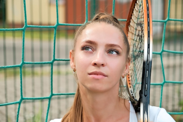 白いスポーツ ドレスを着た女の子が立っているか、近くのテニスコートに座っている テニスコートの女の子の純肖像画