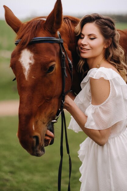 A girl in a white sundress stands next to a brown horse in a field in summer