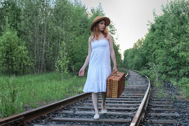 Girl in a white sundress and a wicker suitcase walking on rails