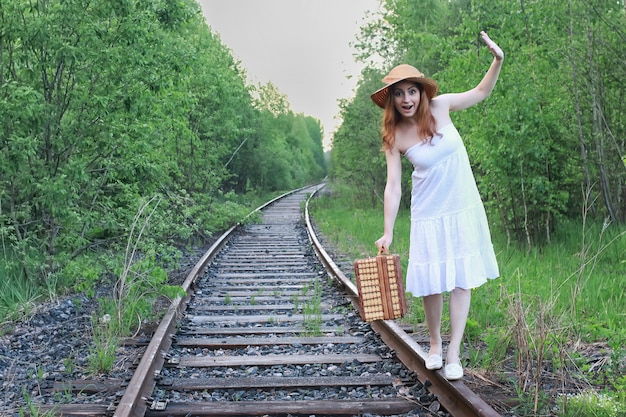 Girl in a white sundress and a wicker suitcase walking on rails