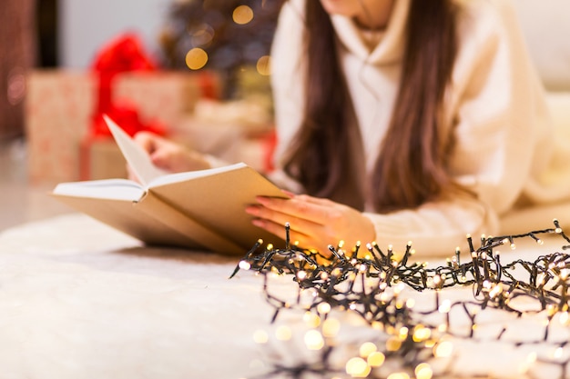 A girl in a white sweater lies and reads a book on a white carpet against the background