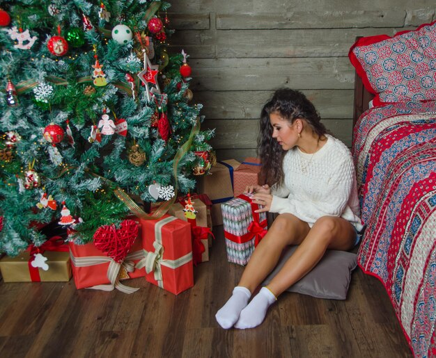 Девушка в белом свитере сидит возле елки и разбирает подарки.