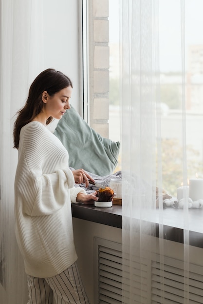 하얀 스웨터를 입은 소녀가 창문 근처에 서서 컵 케이크를 자릅니다.