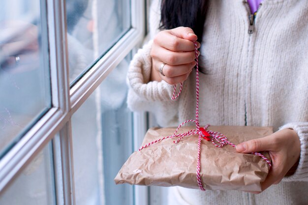 Ragazza in un maglione bianco con un regalo e un candeliere in mano, a casa in salotto vicino alla finestra