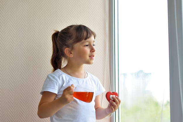 A girl in a white T-shirt holds a cup of tea and a liver in the form of a red heart standing by the window, watching. delicious sweet snack. safe at home, self-isolation during a pandemic.