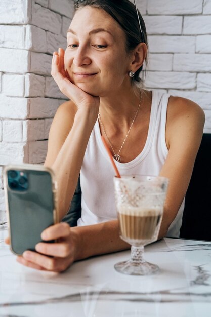 girl in a white t-shirt is talking on the phone via video call in a cafe against a white wall