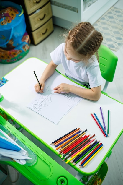 A girl in a white T-shirt sits in her room at the table and draws with colored pencils