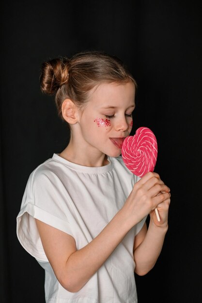 Photo a girl in a white tshirt eats a heartshaped lollipop candy children and sweets baby and sugar