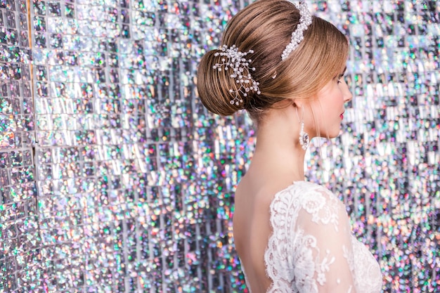 A girl in a white wedding dress with an open back makeup a hairstyle in the form of a bunch poses on silver background mosaic with light spots