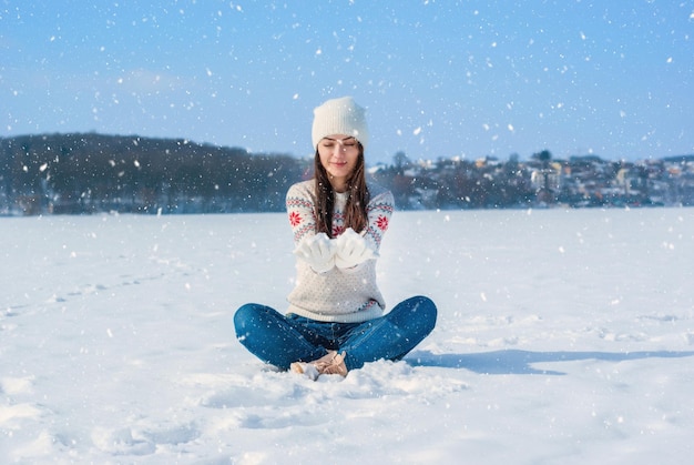 白い冬のセーターを着た女の子 雪の中で足を組んで座っている 雪を上に投げる