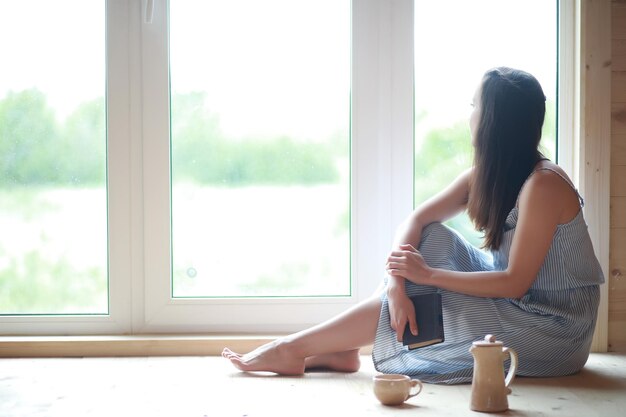 A girl at the window in the house reads a book and drinks tea