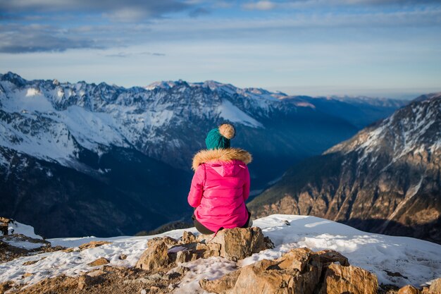 girl in a winter jacket and hat looking at the snowy mountains