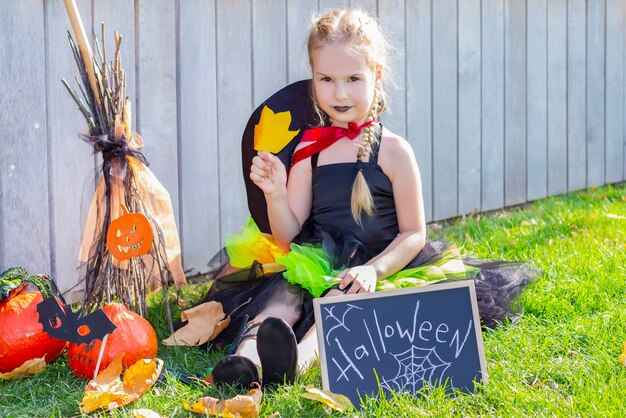 Foto ragazza in costume da strega per le vacanze di halloween. cartello con la scritta: halloween. ragazza seduta sull'erba