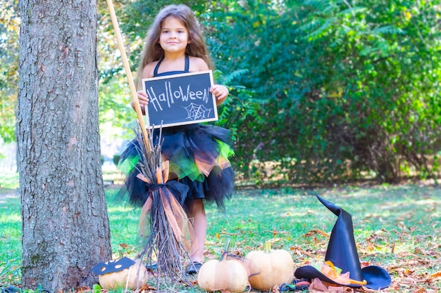 Girl in a witch costume for the Halloween holiday. Placard with the inscription: Halloween. Happy girl standing with a broom