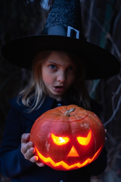ジャコランタンを持った魔女の衣装の帽子をかぶった女の子 大きなカボチャから作られた手作り 目の鼻口のキャンドルライト ハロウィーンの休日のお祝い ナイフで切る野外活動裏庭子供のパーティー