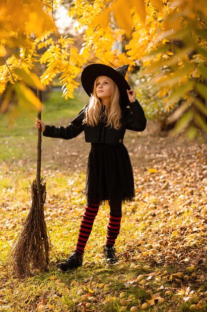 A girl in a witch costume stands with a broom holding onto her hat in an autumn forest