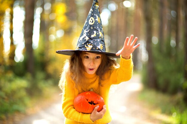 A girl in a witch's hat in a yellow sweater and with a pumpkin with carved eyes and a mouth Jack o Lantern in a yellow autumn forest Halloween Holiday