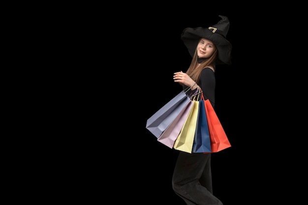 Girl in witchs hat with colorful paper shopping bags in hands is isolated on black background Copy space Black Friday concept