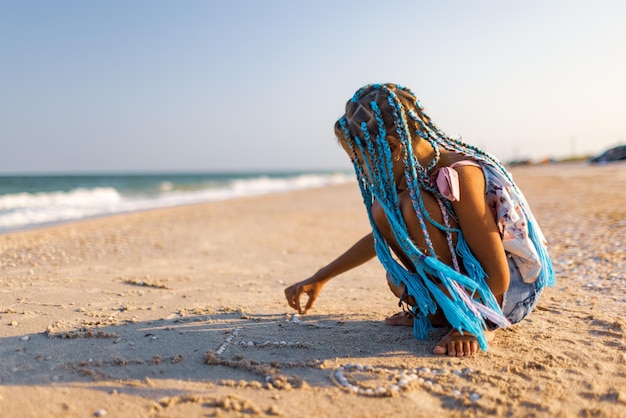 Una ragazza con trecce africane in un costume estivo gioca sulla spiaggia con conchiglie vicino al mare con onde sotto il tramonto soleggiato