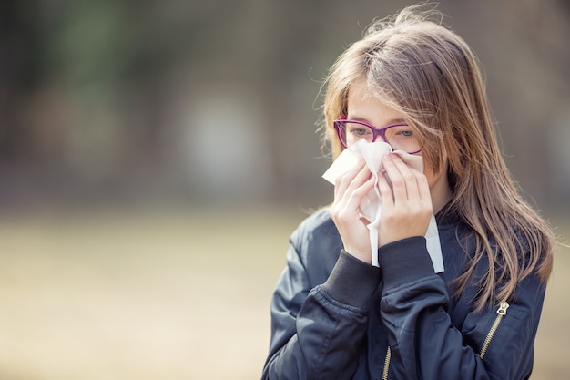 アレルギー症状のある少女が鼻をかむ。公園でティッシュを使用して10代の少女。